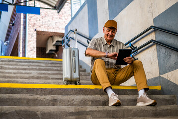 Happy senior man is sitting at railway station and waiting for boarding. He is checking the time on his wristwatch. Senior people traveling.