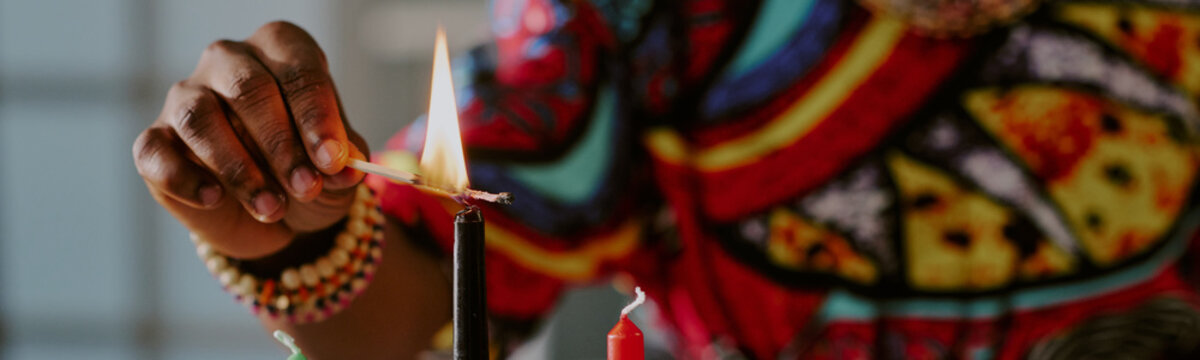 Black woman lighting kinara candle with match during Kwanzaa celebration, hand adorned with colorful beaded bracelets, traditional patterned clothing visible in background