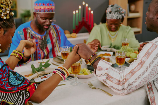 Group of middle aged Black adults sitting at table holding hands and praying during Kwanzaa celebration, traditional kinara with candles and festive food visible in background - Powered by Adobe
