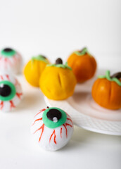 Halloween pastries with small orange and yellow pumpkins and spooky eyeball sweets with red veins. Woman dressed as a witch bites or holds dessert near her eyes on dark textile background.