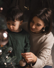 Woman and kid decorate the Christmas tree with baubles, hold presents and sit on a rug near the tree. Mother and son on Christmas or New Year night. Cute boy hangs a globe, cozy semi dark bokeh lights