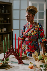 Black woman young adult standing beside kinara with seven candles celebrating Kwanzaa, arranging...