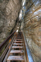 Narrow alpine gorge with wooden walkway, dramatic rock formations and natural light, dark mountain canyon passage creating adventurous hiking atmosphere