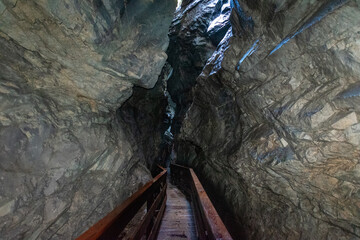 Narrow alpine gorge with wooden walkway, dramatic rock formations and natural light, dark mountain canyon passage creating adventurous hiking atmosphere