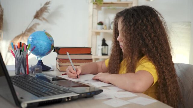 Child studies at desk with laptop while girl writes homework with pencil near globe and book papers on table showing education focus study habit and concentration during home learning session
