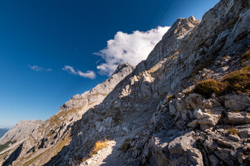 Rugged alpine hiking trail below rocky peaks near the Zugspitze, steep mountain terrain with cable car lines and dramatic high alpine scenery under clear blue sky
