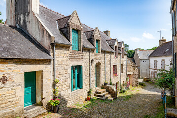 A sunlit cobblestone street winds between ancient stone houses, lush greenery, and blooming plants in Pont-Croix in Brittany, France.