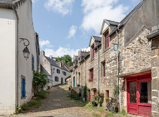 A sunlit cobblestone street winds between ancient stone houses, lush greenery, and blooming plants in Pont-Croix in Brittany, France.