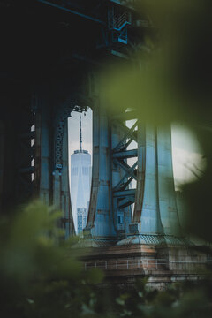 View of the iconic One World Trade Center tower piercing through the steel latticework of the Manhattan Bridge, framed by vibrant green foliage, New York, New York, United States.