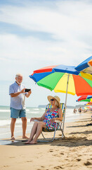 Happy beautiful elderly couple of pensioners. Man taking a picture on smartphone of woman resting under beach umbrellas on the beach, concept of summer vacation for pensioners, hello summer
