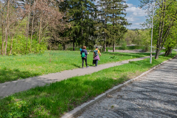 Obraz premium Two people with backpacks walk along a paved path in a vibrant spring park, surrounded by lush green grass and trees under a bright sky. Ideal for themes of nature, leisure, and active lifestyle.