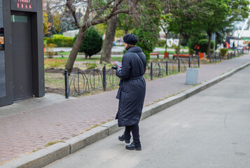 Rear view of a stylish person in a black puffer coat and beret, walking on a city sidewalk while using a smartphone. Urban scene with a public toilet and park in the background. Modern street life.