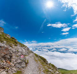 Mountain trail along the Goetheweg above a sea of clouds, narrow alpine path on rocky slope under bright blue sky and sunburst