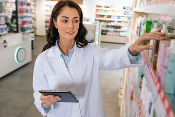 Female worker in white coat writing notes during inspection in pharmacy