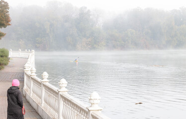 misty atmospheric waterfront nature waterfront along water with kayaking person far on background with walking woman back to camera fall morning season slightly foggy weather