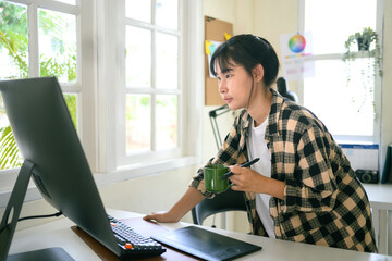Young female graphic designer standing at her desk, holding a coffee cup while working on a computer