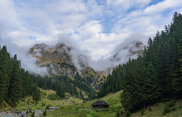 mountain landscape with old cabin of shepherd with two big main peaks in front an vertical forest and rainy clouds on blue sky stana lui burnei