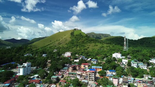 Aerial view across Coron town rooftops with colorful hillside buildings Mount Tapyas green slopes telecom tower and bright daylight clouds over Busuanga Island