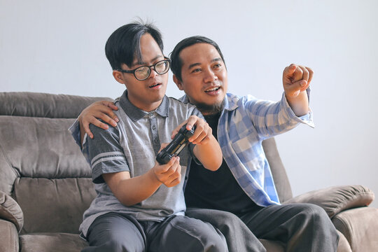 Dad and Son with Special Needs Playing Video Games Together with Joystick on Sofa at Home