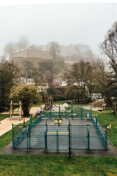 View of an empty playground with a blue fence and green grass, with a building up on a hill covered in fog, Luxembourg city, Luxembourg.