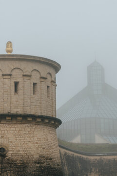 View of a stone tower stands shrouded in mist, a golden finial glinting faintly amidst a glass-roofed building in the distance, Luxembourg city, Luxembourg.