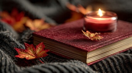 A cozy autumn scene featuring a maroon book, a lit candle, and colorful fallen leaves on a textured blanket.