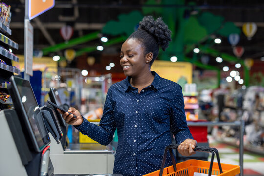 Young black woman scanning groceries and paying with a smartphone at a supermarket self-checkout, smiling while using contactless nfc for fast, convenient retail shopping