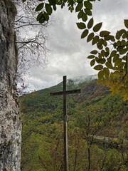 Obraz premium Wooden cross standing on mountain slope with autumn forest in background, cloudy sky with mist and dark clouds above green hills and rocky cliff