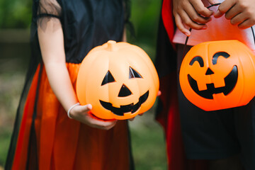 A cheerful young girl dressed in a Halloween witch costume smiles happily outdoors. Wearing a pointed black hat and orange dress, she holds a pumpkin jack-o’-lantern bucket, surrounded by greenery