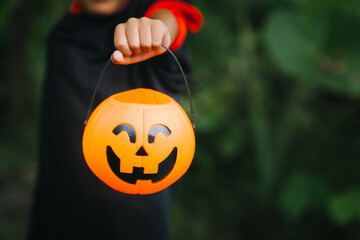 A cheerful young girl dressed in a Halloween witch costume smiles happily outdoors. Wearing a pointed black hat and orange dress, she holds a pumpkin jack-o’-lantern bucket, surrounded by greenery
