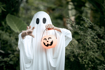 A young boy dressed as a ghost for Halloween wears a white sheet with black eye holes. Holding a...