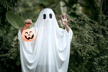A young boy dressed as a ghost for Halloween wears a white sheet with black eye holes. Holding a pumpkin jack-o’-lantern bucket, he stands outdoors surrounded by dark green foliage