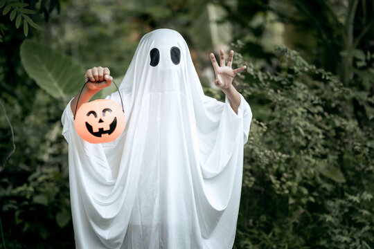 A young boy dressed as a ghost for Halloween wears a white sheet with black eye holes. Holding a pumpkin jack-o&rsquo;-lantern bucket, he stands outdoors surrounded by dark green foliage