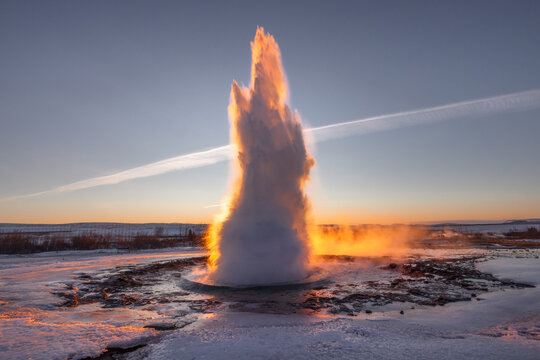 View of a geyser erupts powerfully, illuminated by the warm glow of the setting sun against the icy landscape, creating a dramatic contrast, Iceland.