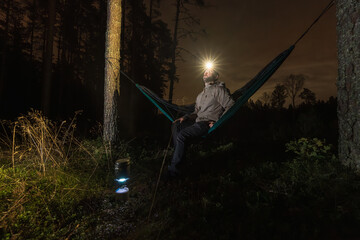 Hiker with a headlamp prepares a hot tea on a camping stove while resting in a hammock at night. © Dmitri