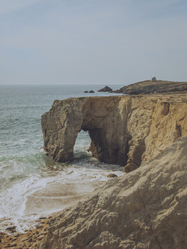 View of the  golden cliffs  of Arche De Port Blanc Roche Perc&Atilde;&copy;e arching over turquoise waves crashing on the shore, a distant lighthouse standing sentinel, Saint-Pierre-Quiberon, Brittany, France.