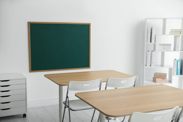 Blank green chalkboard, desks, chairs and shelving unit in classroom