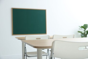 Blank green chalkboard, desks, chairs and plant in classroom, closeup