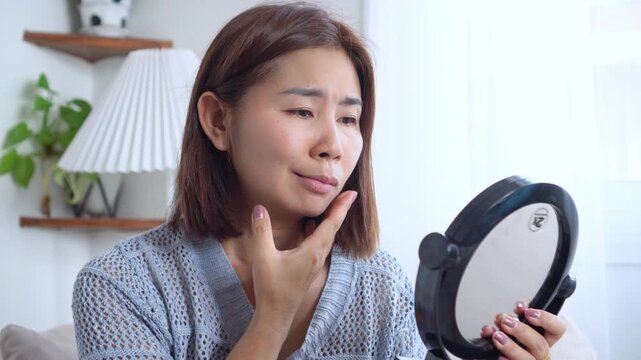 Asian woman suffering from facial palsy, depressed mood, drooping mouth and hand on face while looking in the mirror