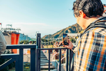A young Hispanic man with dark hair takes a photo with his smartphone while standing on a balcony overlooking a mountain landscape and recreational area.