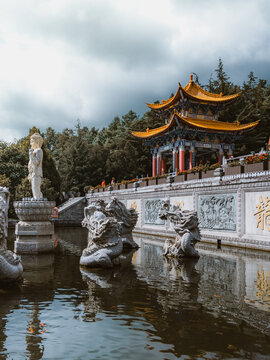 View of a tranquil pond reflecting ornate stone sculptures and a traditional Chinese pagoda under a cloudy sky, Dali, Yunnan, China.