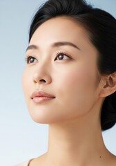 Fototapeta premium Closeup portrait of a young asian woman with clear, glowing skin, looking thoughtfully to the side against a soft blue background, highlighting natural beauty and skincare