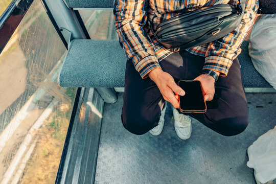 A young man with dark hair and a plaid shirt sits in a cable car, using a smartphone. The scene captures a vacation atmosphere in the mountains.