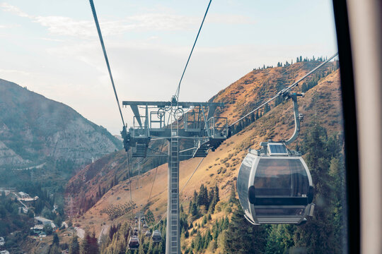 A cable car travels up a mountain slope. The landscape features rocky terrain and trees. The sky is clear with soft clouds, indicating a pleasant vacation atmosphere.
