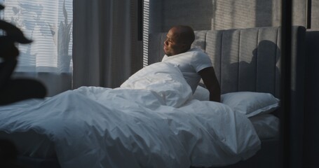 In Modern Bedroom, Adult Man Wakes Up in White Bed. Sleepy Guy Squints Eyes From Bright Light and Gets Out of Bed, Stretching. Concept of Healthy Sleep, Relaxation, and Morning Routine. Static Shot.