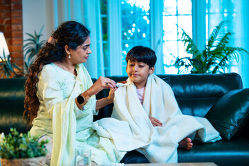 Obraz premium Indian Mother taking son’s temperature while sitting on sofa, showing love and parental care