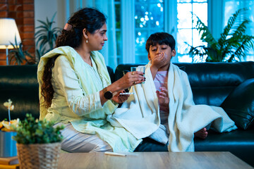 Indian Mother taking son’s temperature while sitting on sofa, showing love and parental care