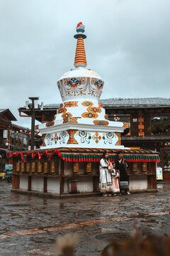 View of a gleaming white stupa adorned with intricate patterns stands majestically amid the old town's grey stone, where a couple embraces, Di Qing Zang Zu Zi Zhi Zhou, Yun Nan Sheng, China.