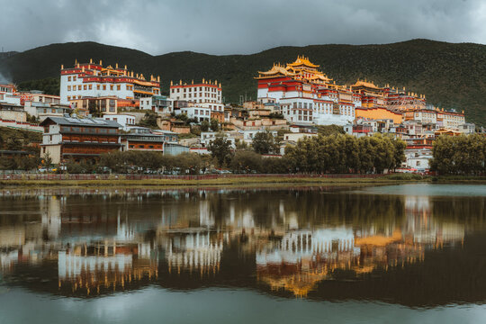 View of golden roofs and red walls cascade down the hillside, mirrored in the still waters below, creating a serene tapestry of color and reflection, Di Qing Zang Zu Zi Zhi Zhou, Yun Nan Sheng, China.