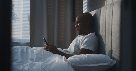 Happy African American Man Lies Comfortably in Modern Bedroom, Using Smartphone While Leaning on...